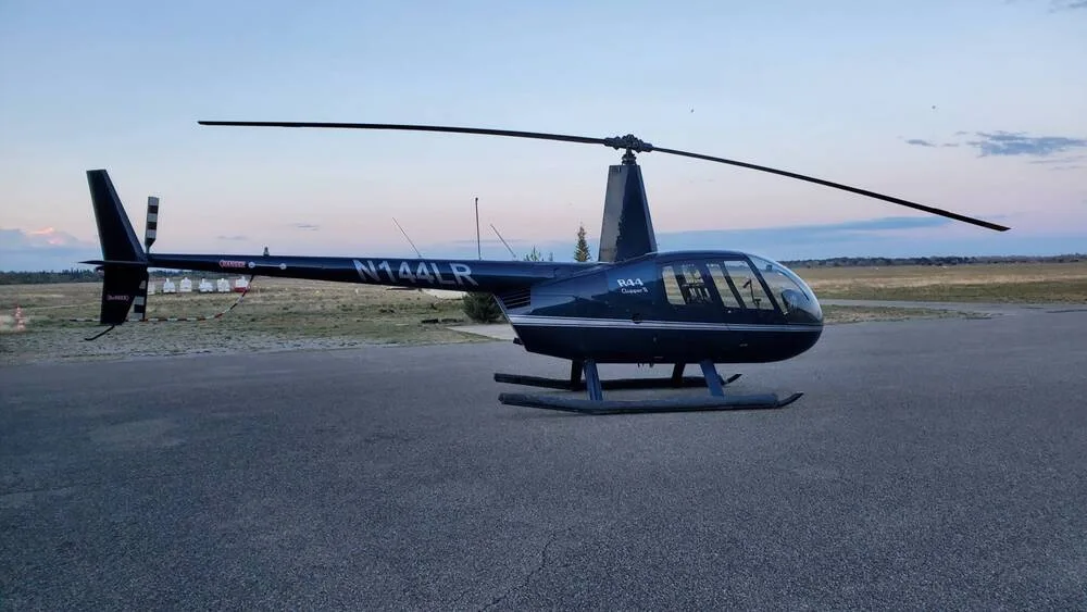 A dark blue Robinson R44 Clipper II helicopter, N144LR, sits on an asphalt pad at sunset.