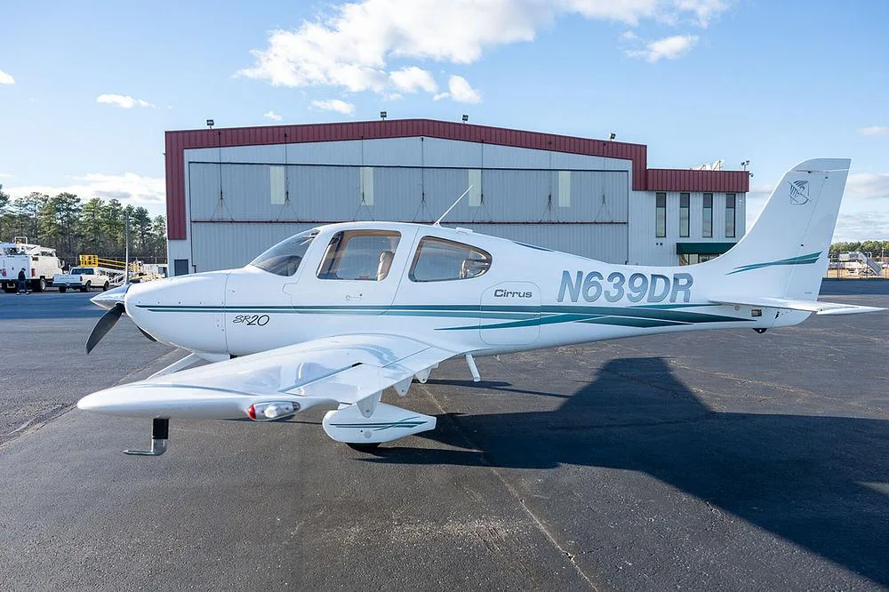 A white Cirrus SR20 aircraft with green and grey stripes, registration N639DR, parked on an asphalt tarmac in front of a hangar.
