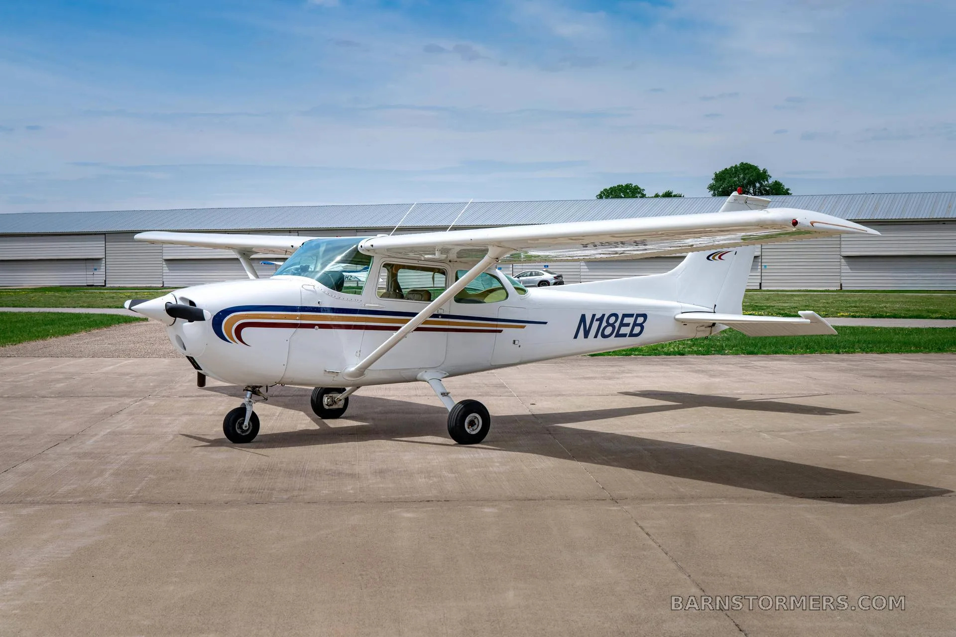 A white Cessna 172 (N18EB) with blue and gold stripes is parked on a tarmac, viewed from the front-right.