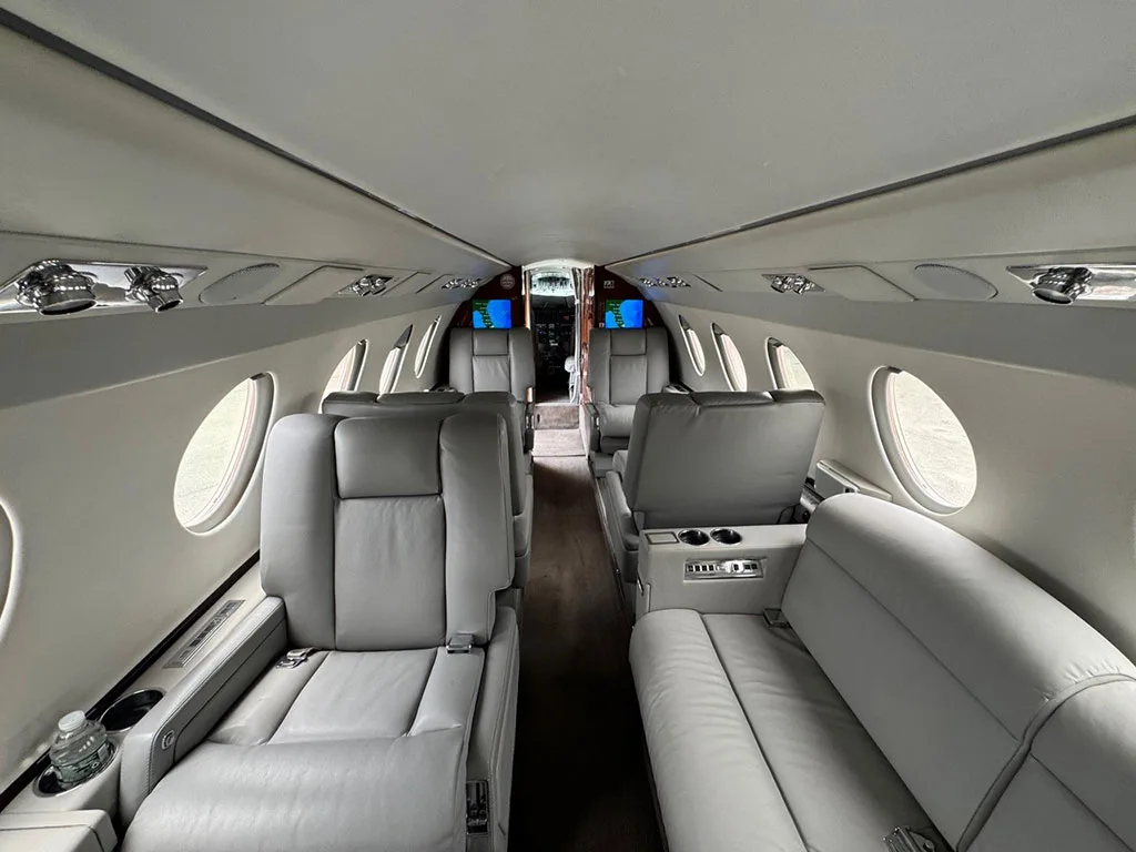 View down the aisle of a business jet cabin with light gray leather seats and wood accents, looking towards the cockpit.