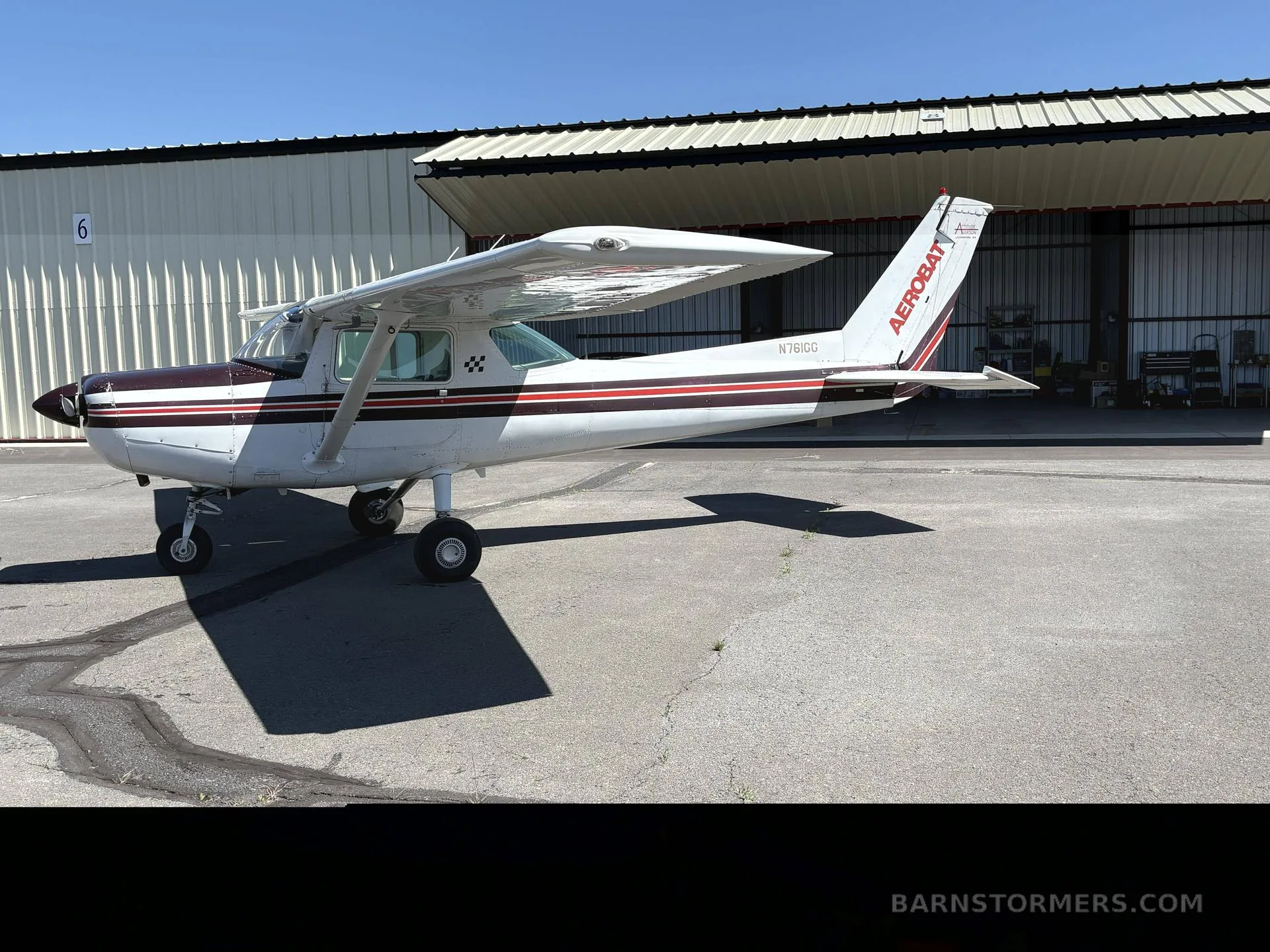 A white and maroon Cessna 150 Aerobat (N761GG) parked on an asphalt apron next to a hangar.