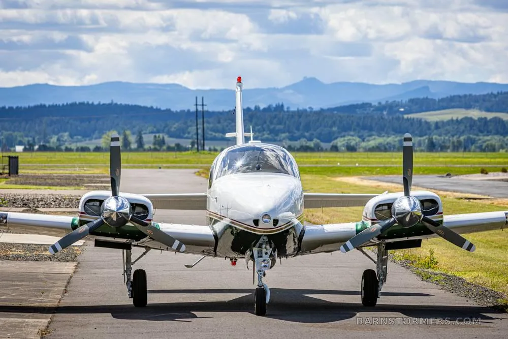 A white and green twin-engine propeller aircraft is viewed head-on on a tarmac with distant mountains under a cloudy sky.