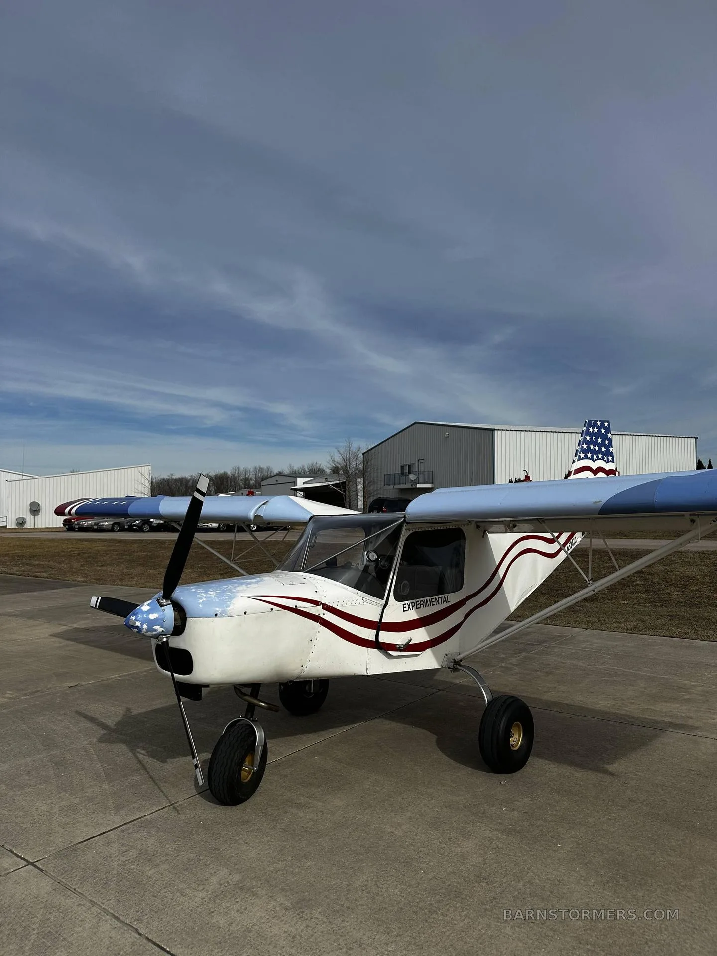 A white and blue experimental aircraft with a red and white striped tail and fuselage, parked on a concrete ramp in front of hangars under a cloudy sky.