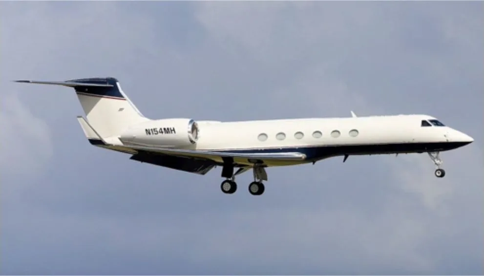 A white Gulfstream G550 business jet with a dark blue stripe, registration N154MH, in flight with landing gear extended against a cloudy sky.