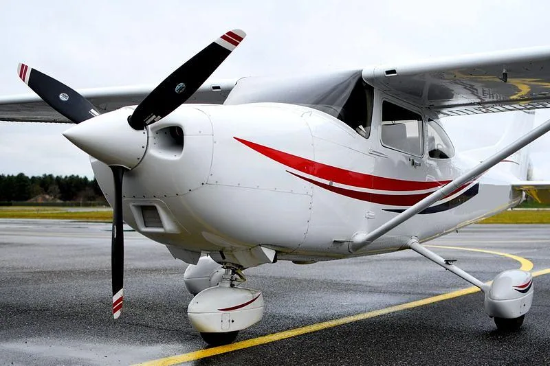 A white single-engine propeller aircraft with red stripes on the fuselage is parked on a wet tarmac under an overcast sky.