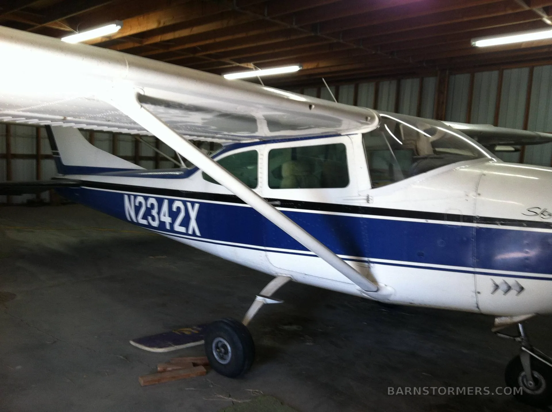 A white and blue Cessna 182 Skylane (N2342X) parked inside a hangar with wooden ceiling beams.