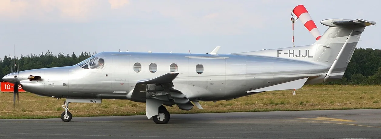 A silver Pilatus PC-12 aircraft with registration F-HJJL taxiing on an airfield, viewed from the side with a red and white windsock in the background.