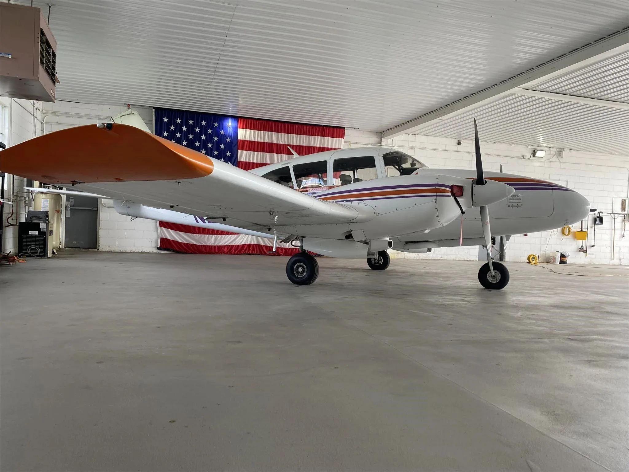 A white twin-engine Piper Aztec aircraft with orange and purple stripes parked in a hangar with an American flag in the background.