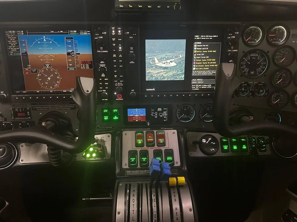 The cockpit of an aircraft features multiple digital displays, analog gauges, and illuminated switches, with a small image of an aircraft on one screen.