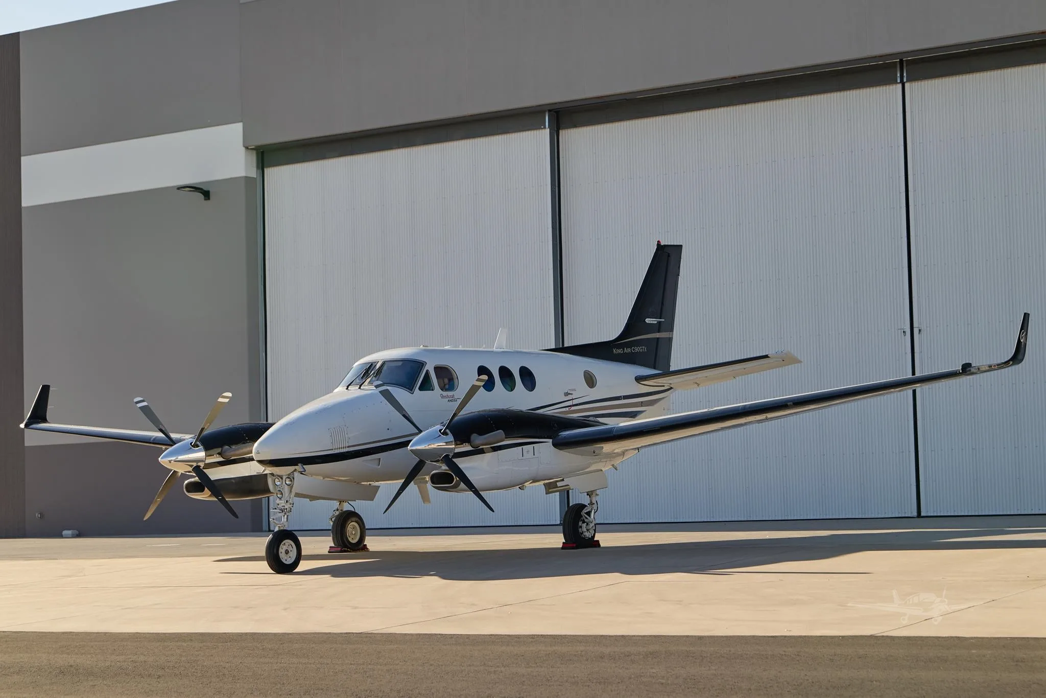 A white and black Beechcraft King Air C90GTx turboprop aircraft is parked on a tarmac in front of a hangar.