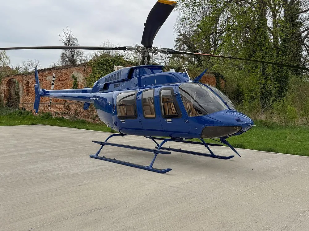 A blue Bell 505 helicopter is parked on a concrete pad next to an old brick building and trees.