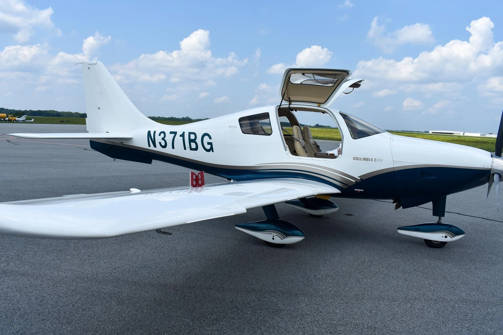 A side view of the white and blue Columbia 400 aircraft, registration N371BG, with its gull-wing doors open, parked on an airfield.