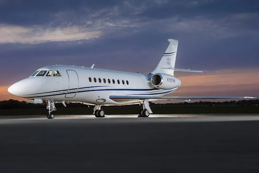 A white Dassault Falcon 900EX private jet with blue stripes and tail number N192HA, parked on an airfield at dusk.
