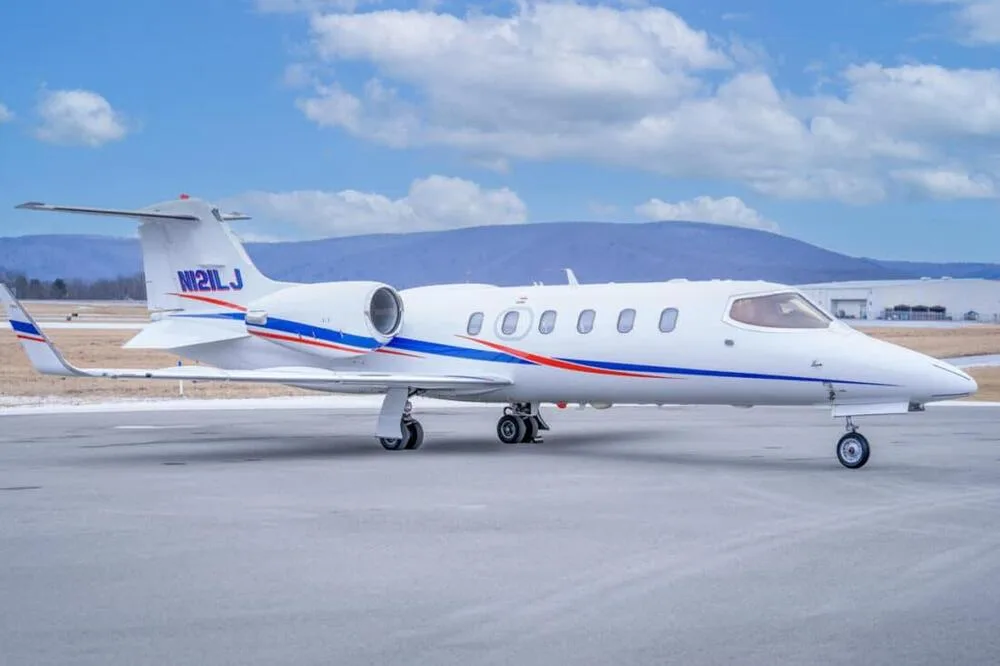A side view of a white Learjet 45 with blue and red stripes and registration N121LJ, parked on a tarmac with mountains in the background.