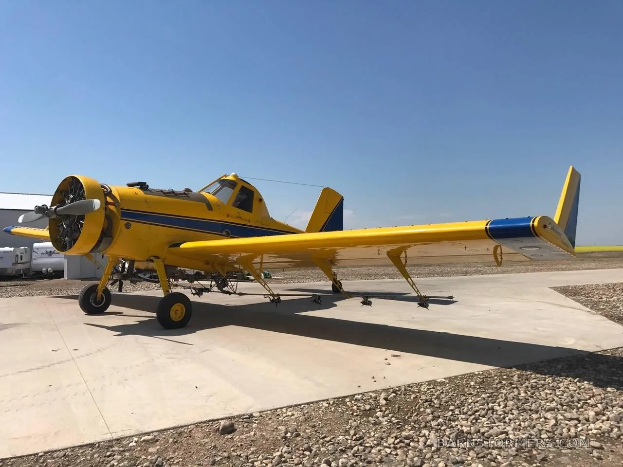 A yellow agricultural aircraft with blue stripes parked on a concrete pad next to a gravel area under a clear blue sky.