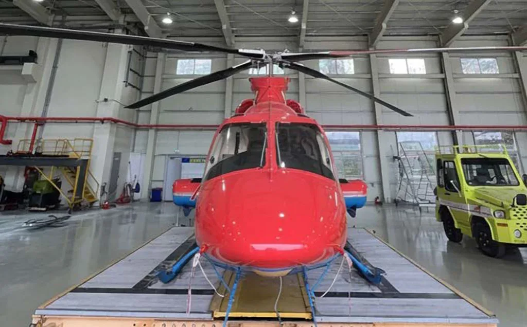 A red and blue helicopter is viewed from the front, sitting on a platform inside a hangar.
