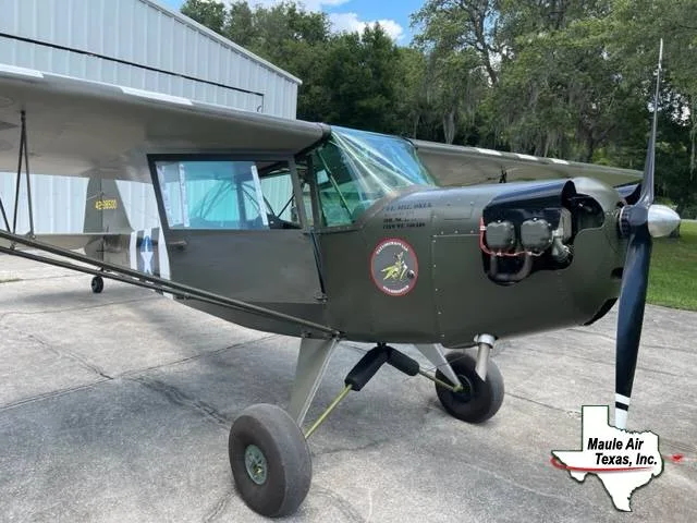 A military green, single-engine, high-wing aircraft with a visible engine and a white star insignia on the tail, parked on concrete next to a hangar.