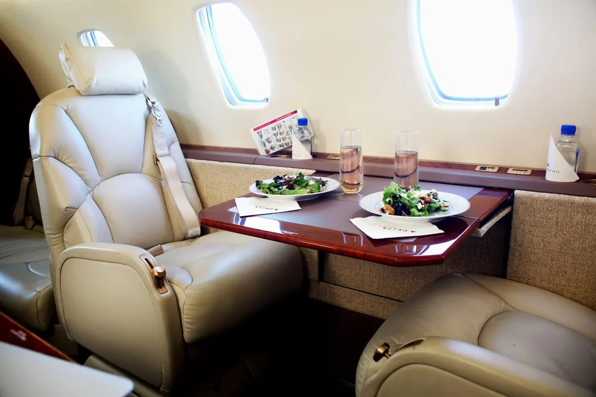 The interior of a private jet cabin features two beige leather seats and a table set with salads and drinks.