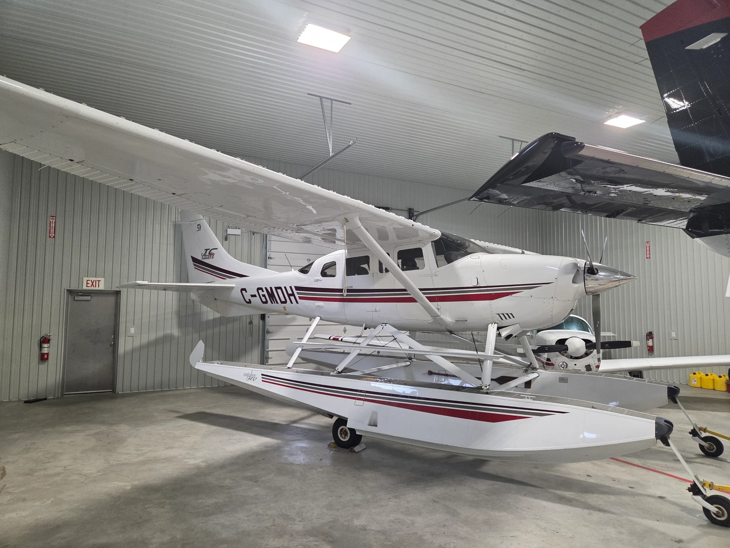 A white Cessna 206 Stationair floatplane with red and black stripes is parked inside a hangar.