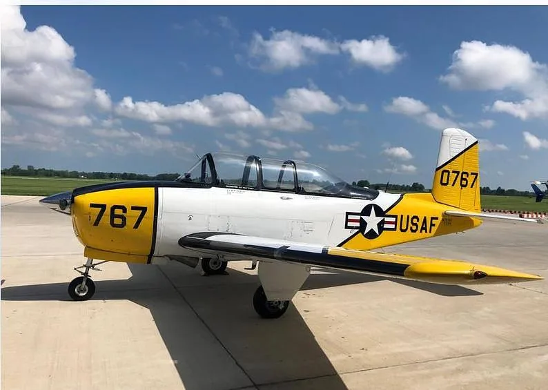 A white and yellow T-34 Mentor aircraft with "USAF" markings and "767" on the tail and nose, parked on an airfield under a partly cloudy sky.
