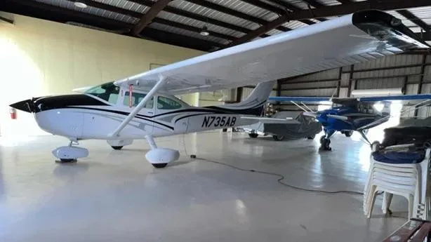 The white Cessna aircraft N735AB with black and grey stripes, parked in a hangar with other aircraft in the background.