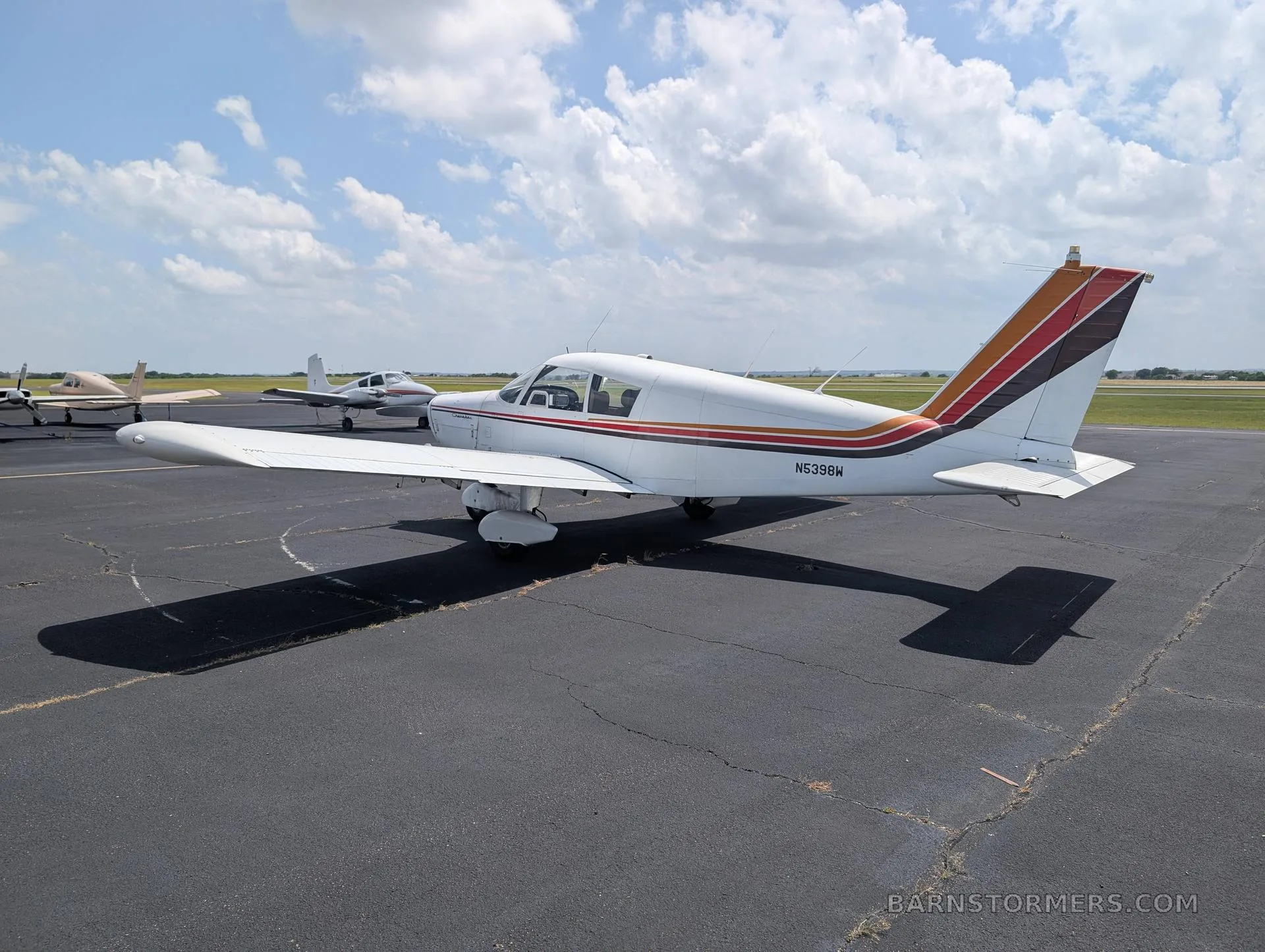 A white single-engine low-wing aircraft with red, orange, and brown stripes on the tail and fuselage, viewed from the rear-right, parked on an asphalt ramp.