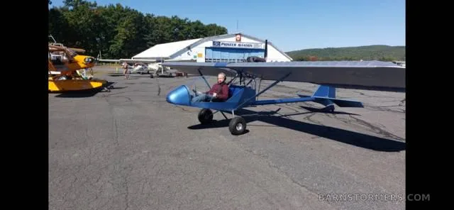 A blue ultralight aircraft with a pilot sitting in it, parked on an airfield in front of a hangar.