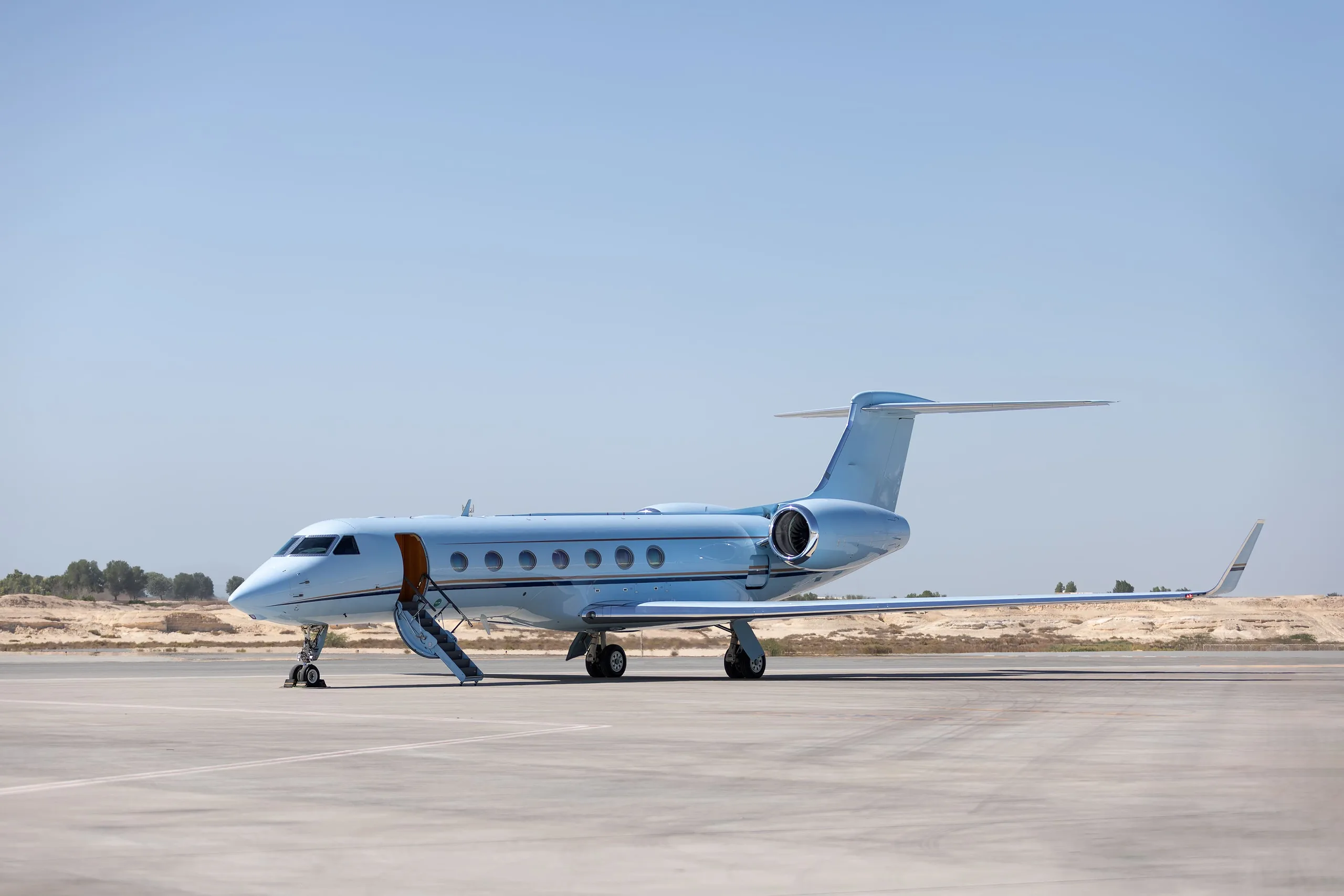 A light blue Gulfstream business jet with an open door and stairs extended, parked on a tarmac in a desert-like setting.