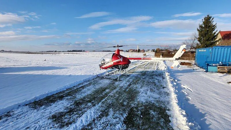 A red and white Kompress helicopter with registration OK-KHA11 is parked on a cleared path in a snowy field under a clear blue sky.