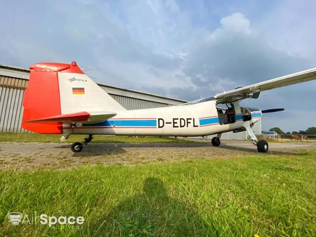 A white aircraft with blue and red stripes and a red tail, parked on a grassy field next to a hangar.