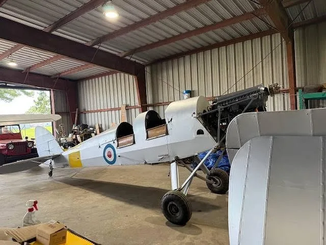 A white biplane with a yellow stripe and roundel, partially assembled, sits in a hangar.