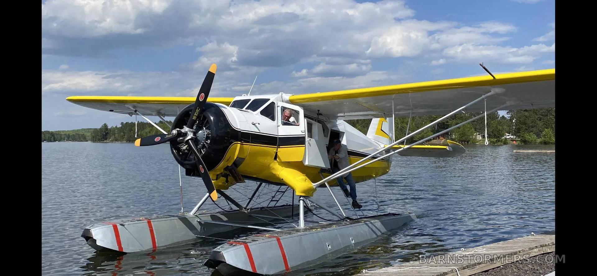 A yellow and white floatplane is partially in the water, with a person entering the cabin, under a cloudy sky.