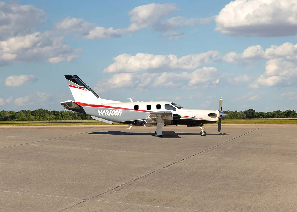 A white and grey TBM 900 series aircraft with red stripes, registration N180MF, parked on an airfield, viewed from the front-right.