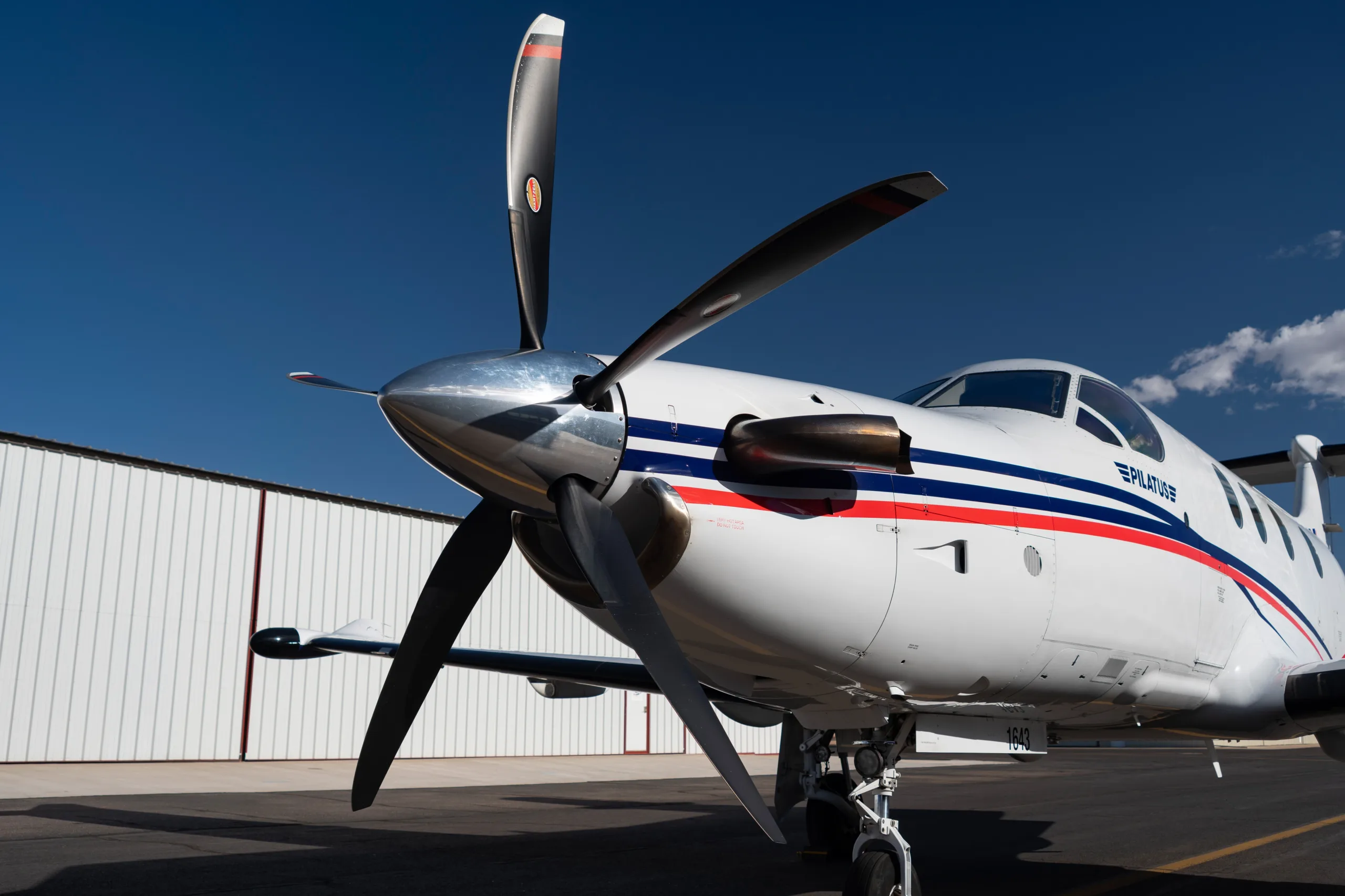 A close-up view of the propeller and nose of the Pilatus PC-12 aircraft, N985AW, with a hangar in the background.