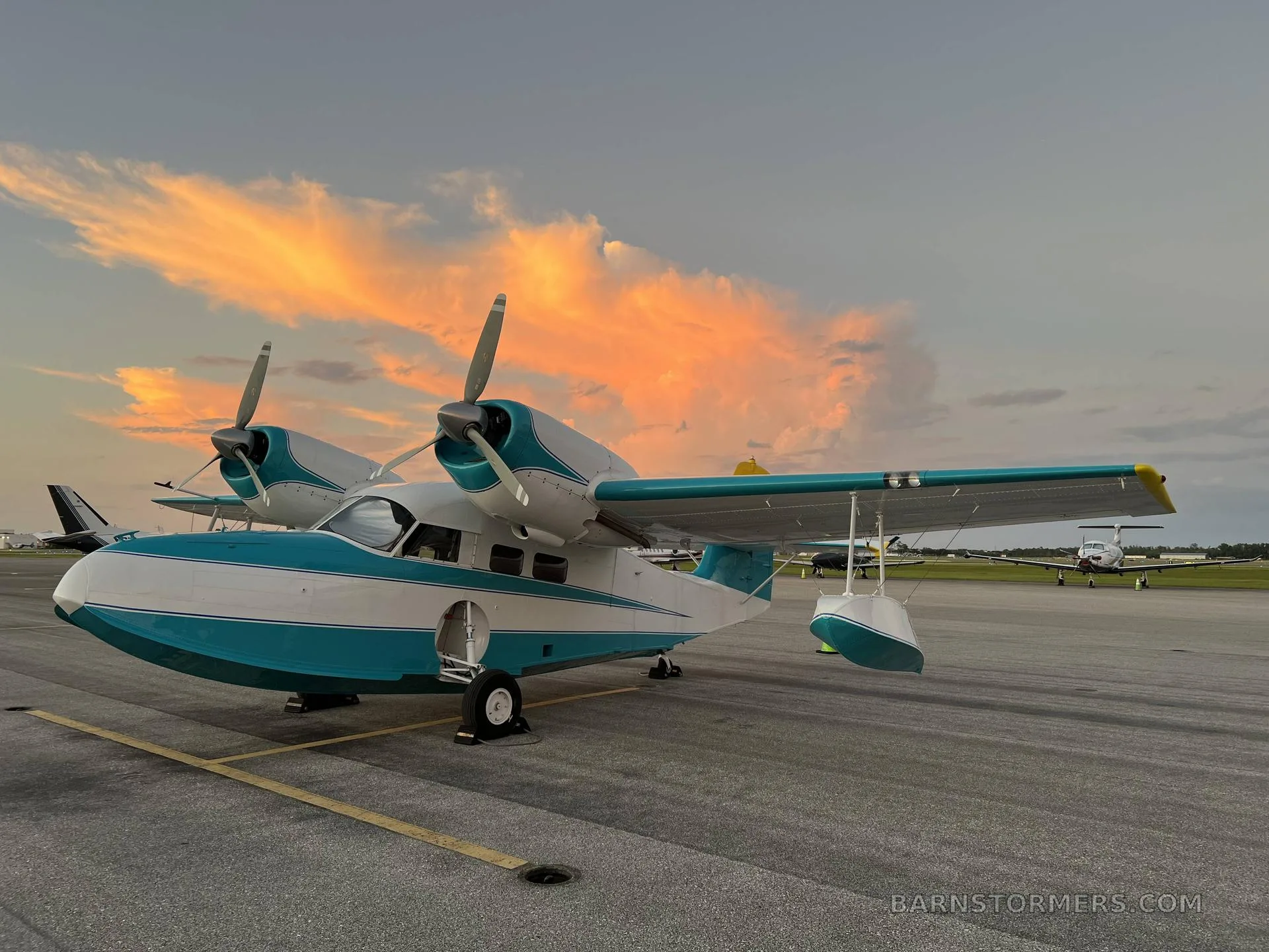 A white and teal Grumman G-44 Widgeon seaplane is parked on an airfield tarmac at sunset, with other aircraft in the background.