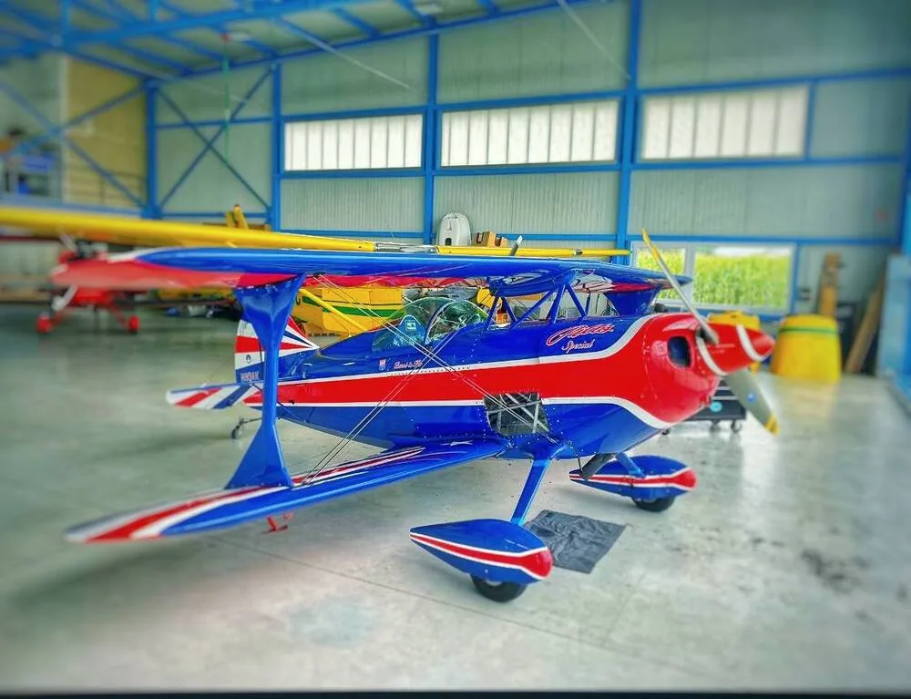 A red, white, and blue Pitts Special biplane, N99AK, is parked inside a hangar.