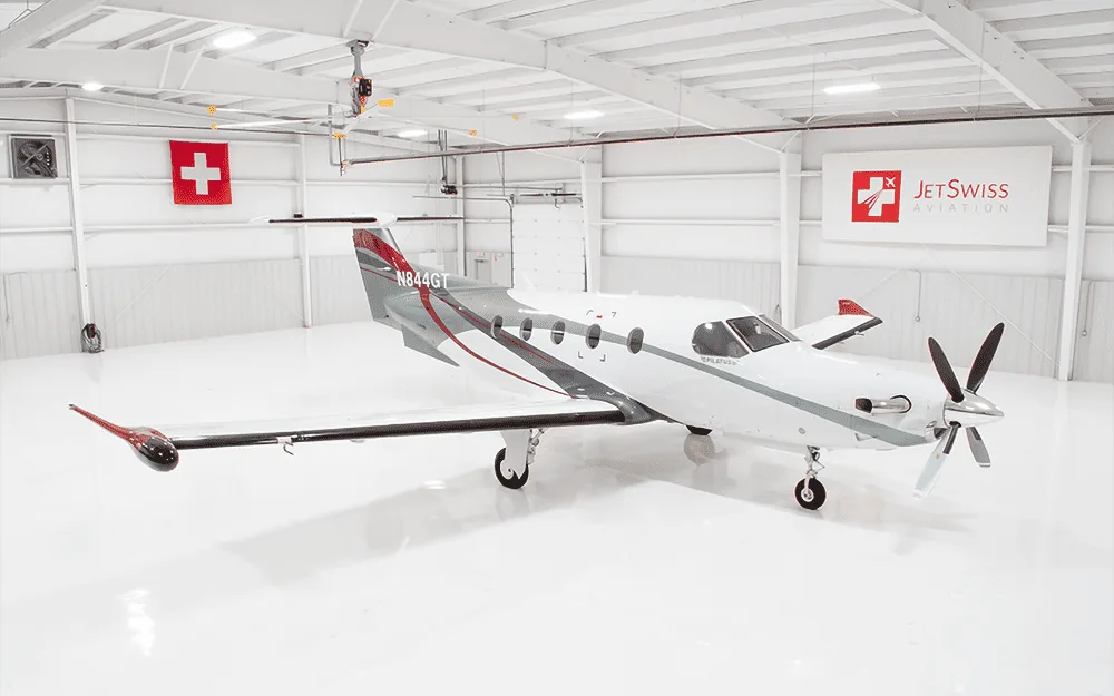 A white Pilatus PC-12 NGX aircraft with red and grey accents is parked in a brightly lit hangar.