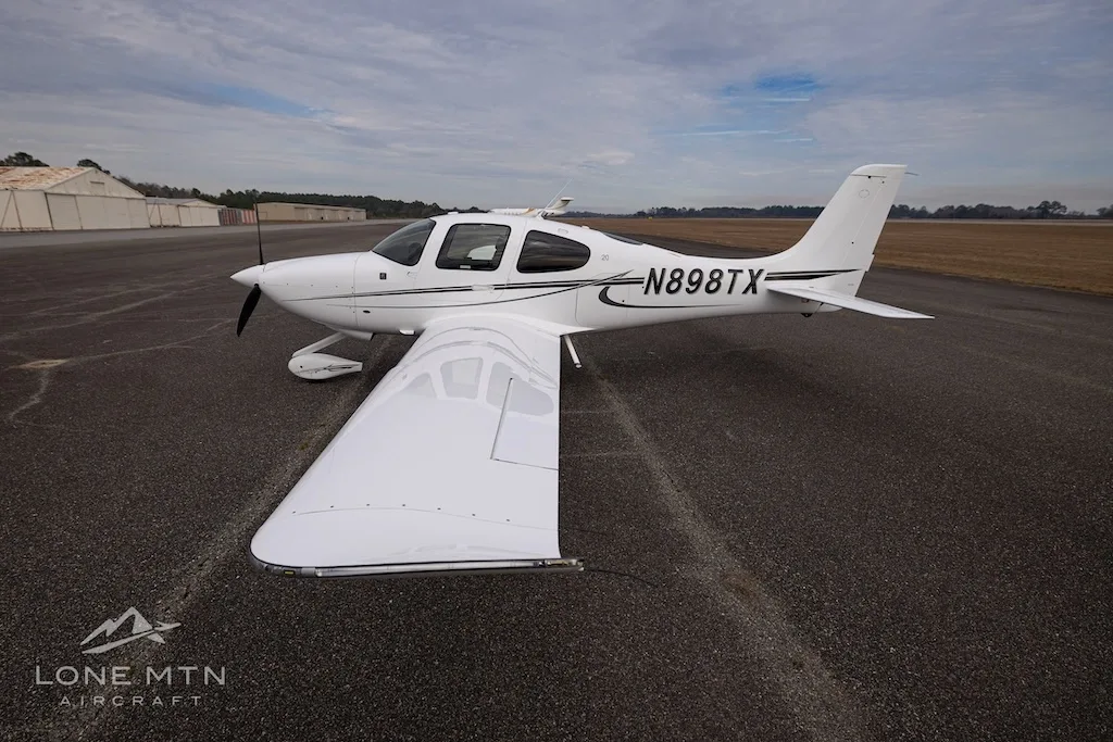 A white Cirrus SR20 aircraft with black and grey stripes, registration N898TX, viewed from above and to the right on an asphalt runway.