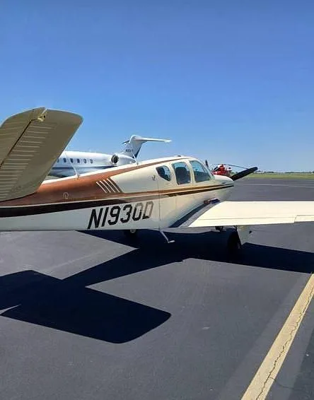 The tail section of a white and brown single-engine aircraft, N1930D, parked on an airfield with other aircraft in the background.