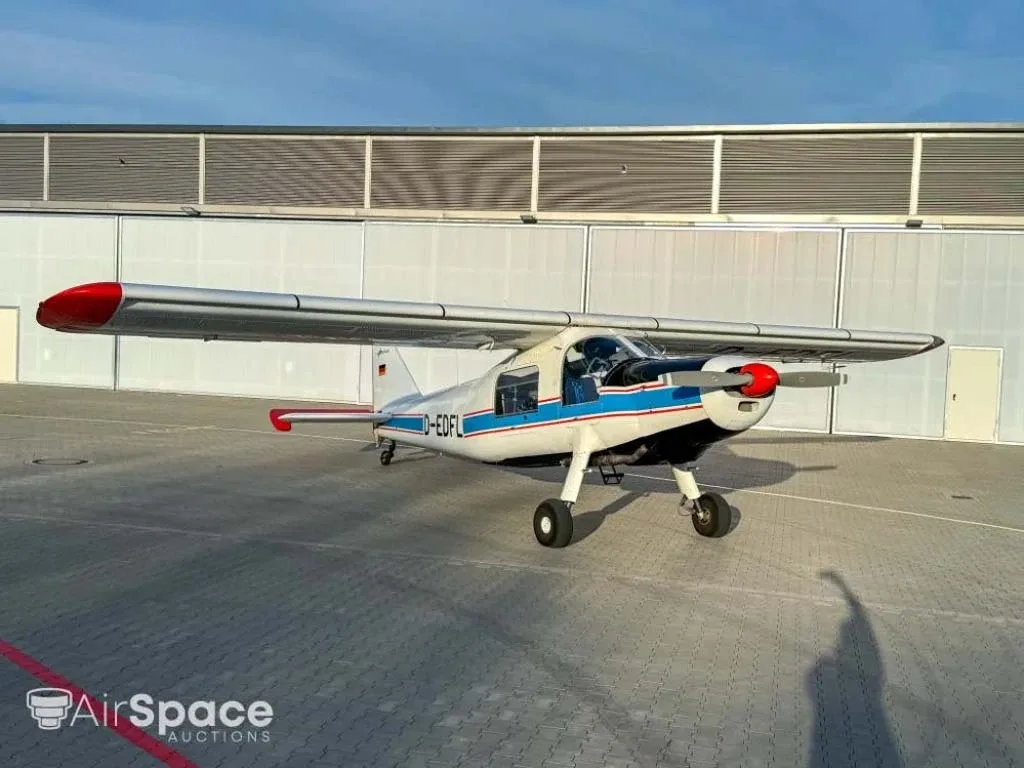 A white aircraft with blue and red stripes and red wingtips, parked on a paved apron in front of a hangar.