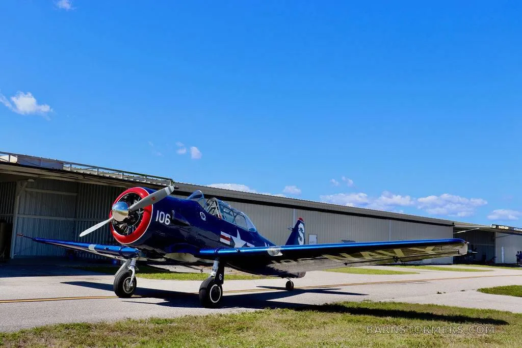 A dark blue vintage military trainer aircraft with a red nose and white propeller is parked on an asphalt ramp next to hangars under a clear blue sky.