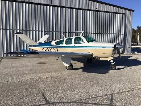 A beige and blue single-engine aircraft with registration C-GXOZ is parked on an asphalt apron next to a corrugated metal hangar.
