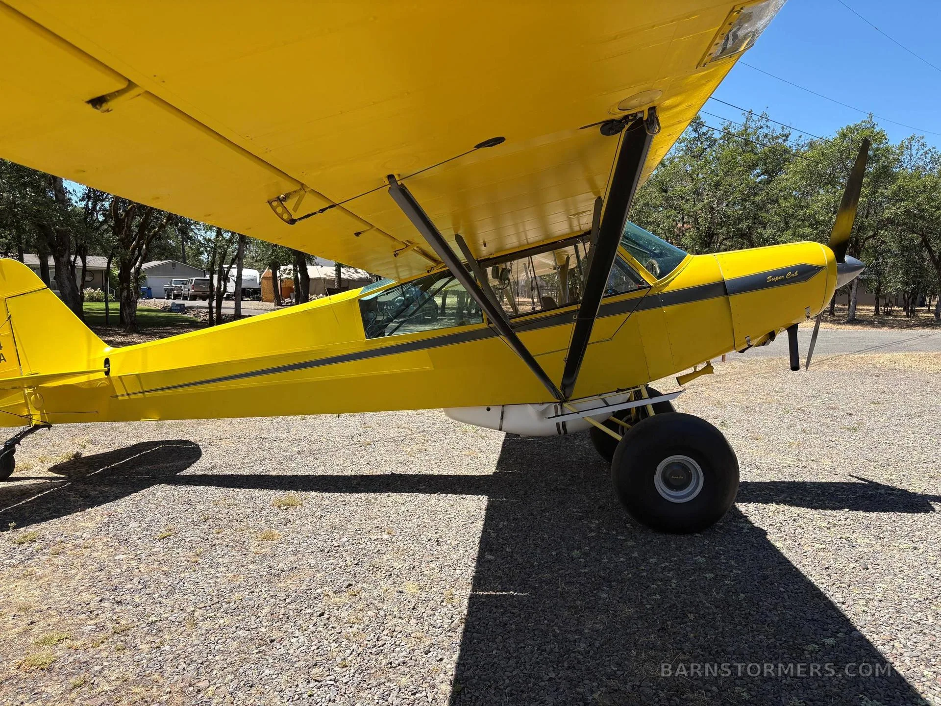 A side view of a yellow Piper Super Cub with black stripes and large tundra tires, parked on a gravel surface.