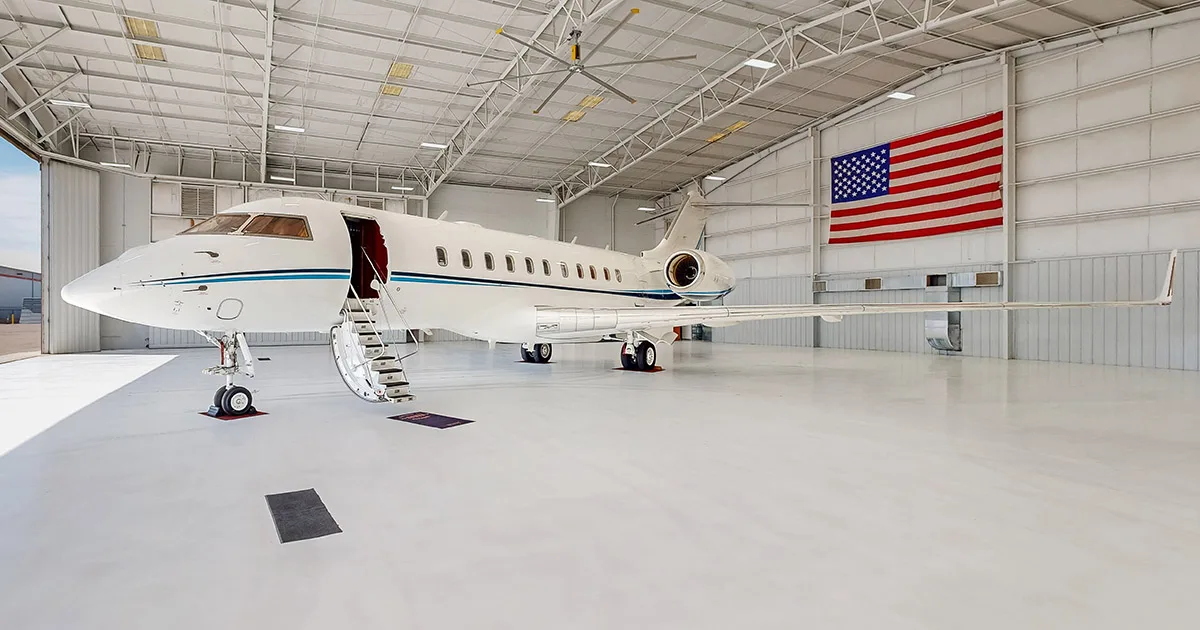 A white business jet with blue stripes parked in a brightly lit hangar with an American flag on the wall.