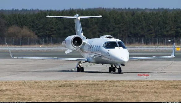 A white business jet is seen from a front-quarter view on an airport tarmac with trees in the background.