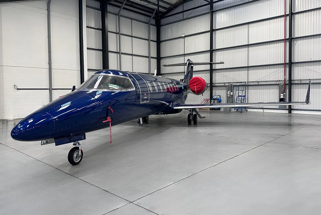 A dark blue private jet with red engine covers parked inside a hangar, viewed from the front-right.