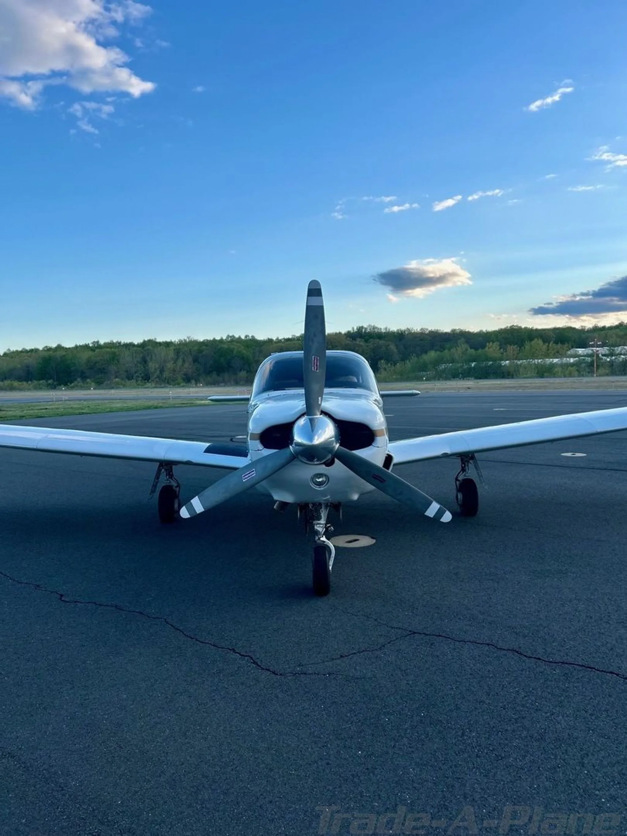 A front view of a white Piper Cherokee aircraft with a propeller, parked on an airfield under a blue sky.