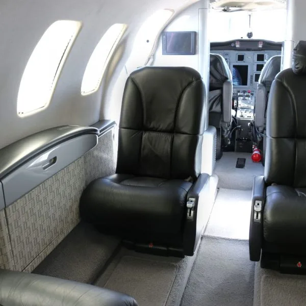 A side view of a black leather seat in a private jet cabin, with the cockpit visible in the background.