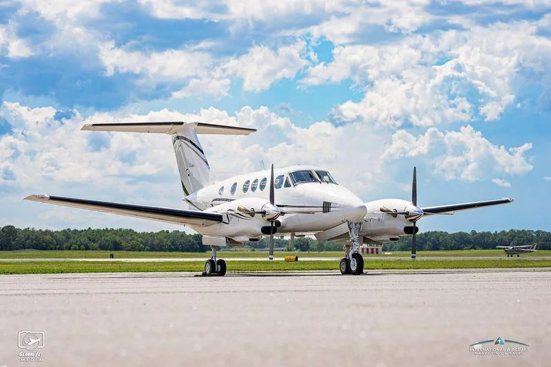 Three-quarter front view of a white twin-engine turboprop aircraft with black and gold accents parked on a tarmac.
