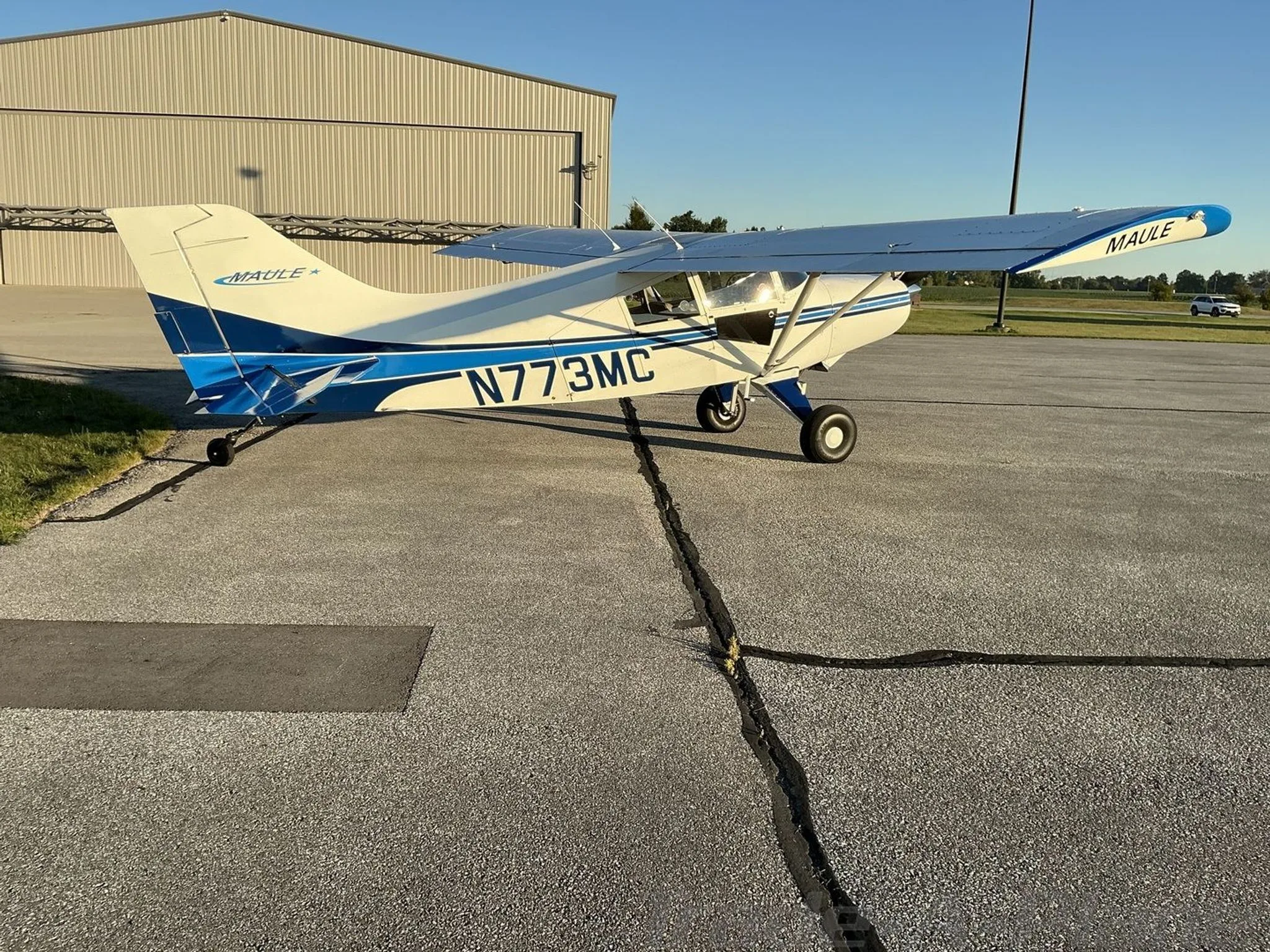 A white Maule aircraft with blue tail and wingtips, parked on an airfield next to a hangar.